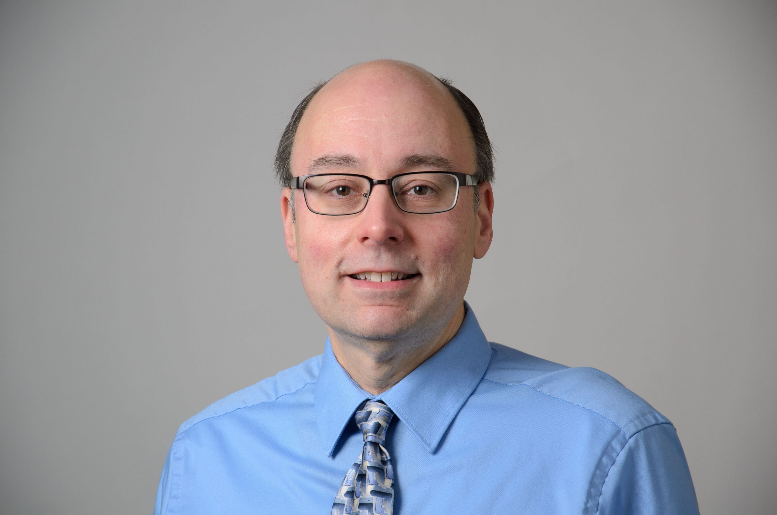 Headshot of David Cavallaro. blue shirt, and patterned tie, black glass frames.